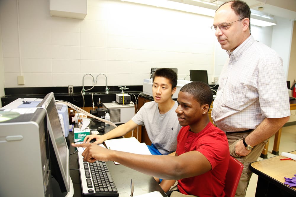 two students working on a computer with a teacher standing behind them