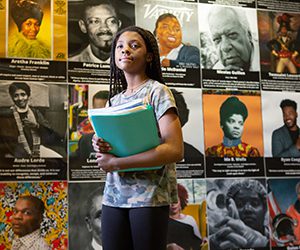 A black girl standing holding a notebook with a backdrop of images of civil rights heroes behind her