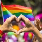 Two individuals forming heart with hands in front of pride flag