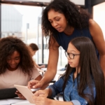 Teacher of color assisting two girl students of color in a classroom