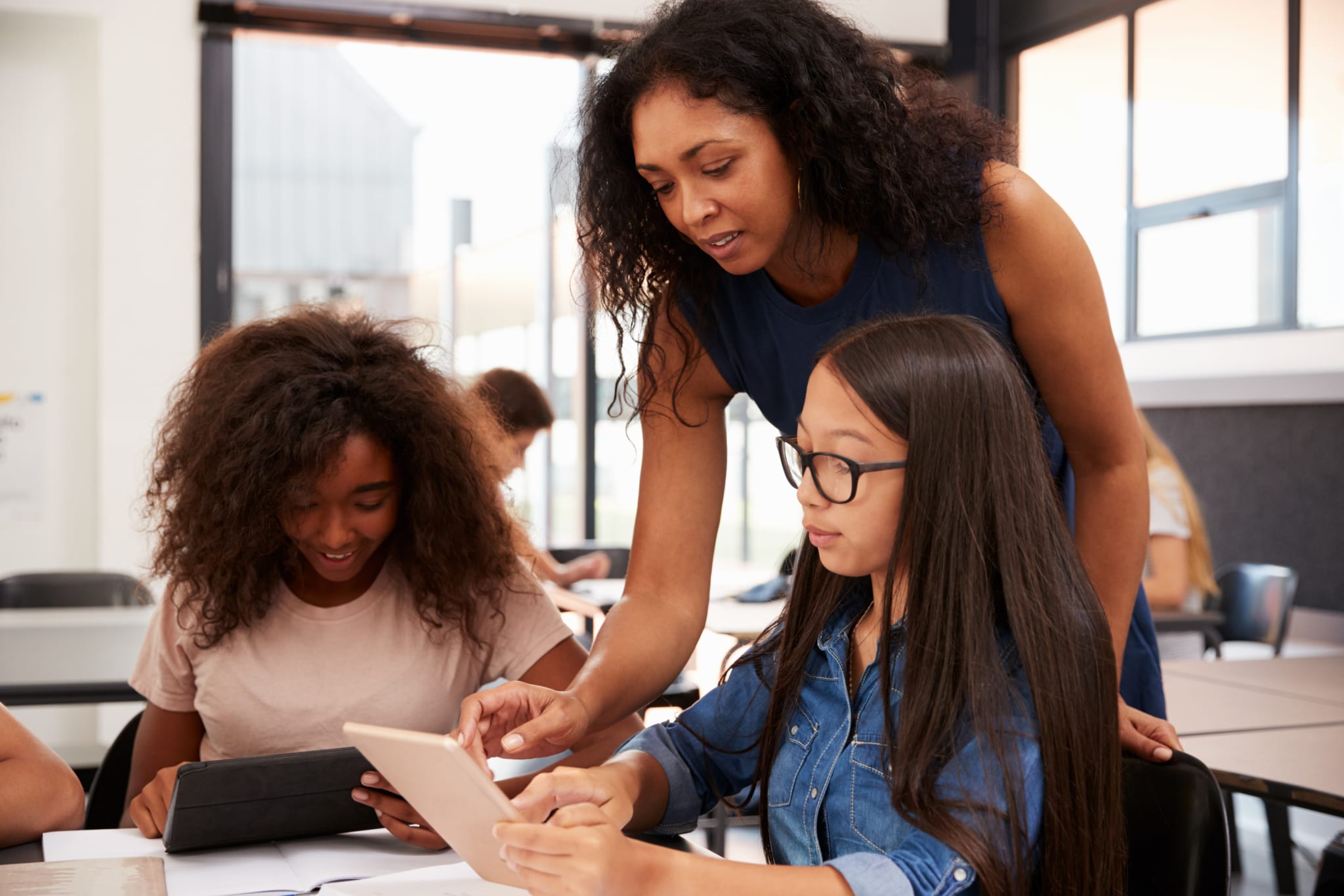 Teacher of color assisting two girl students of color in a classroom