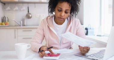 Black woman working on calculator and laptop and holding paper in hand