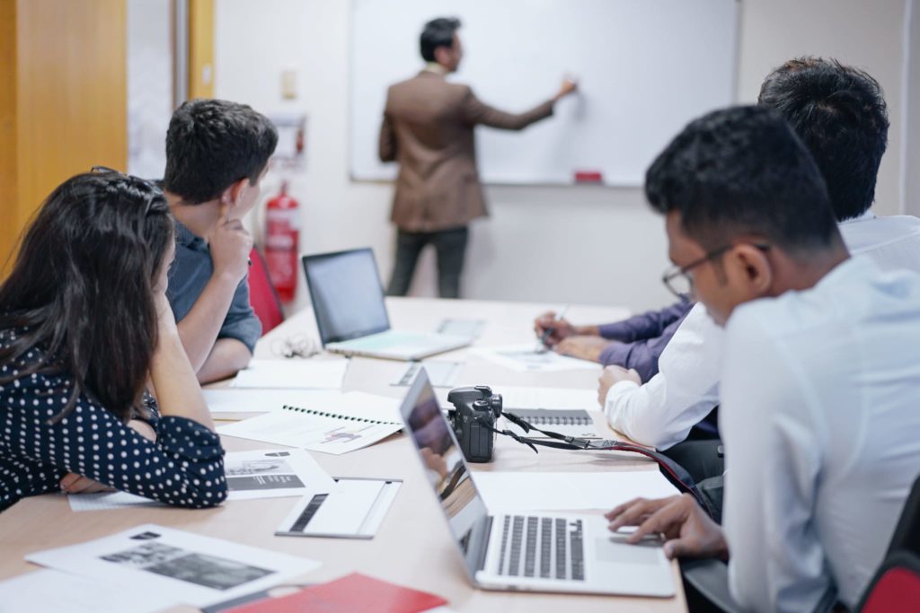 A teacher writing on a whiteboard as students watch sitting down