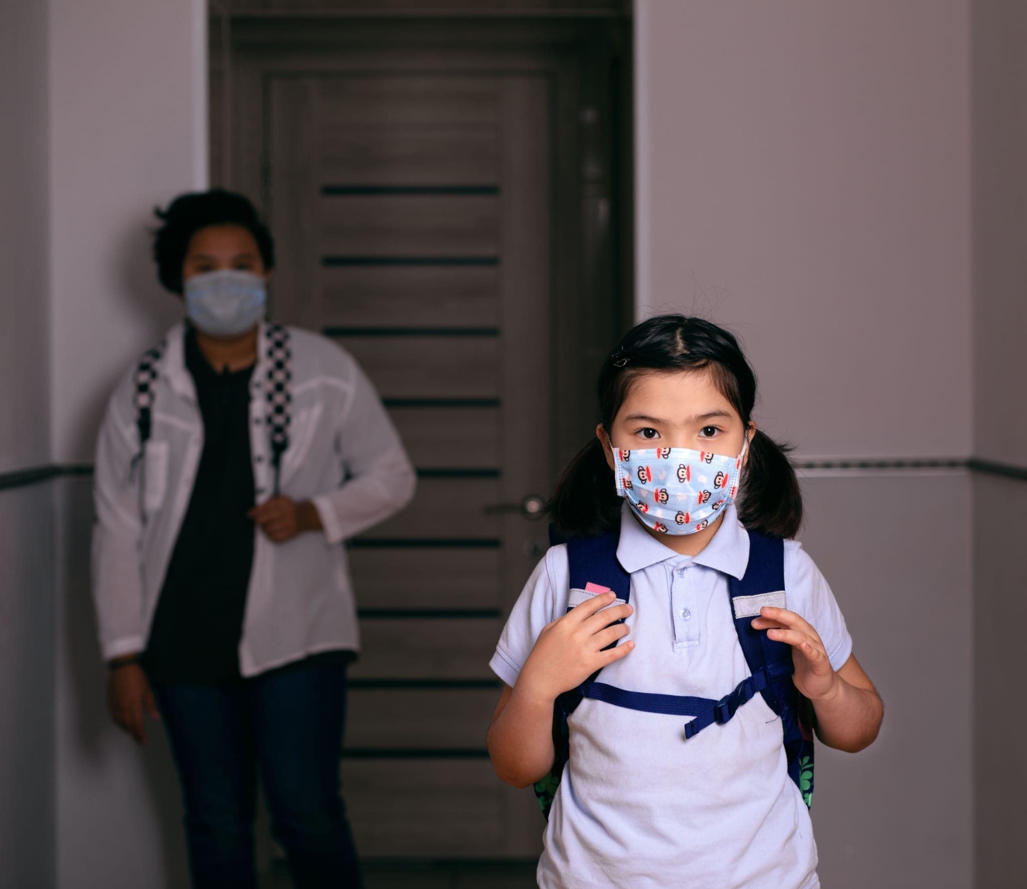Child wearing a face mask and backpack while mother stands behind her with a face mask on