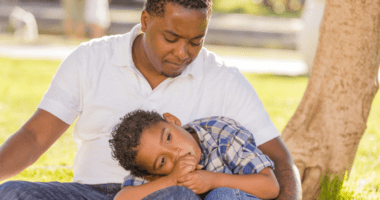 African American man sitting on the ground near a tree with his young son laying his head on his lap