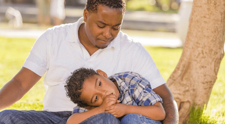 African American man sitting on the ground near a tree with his young son laying his head on his lap