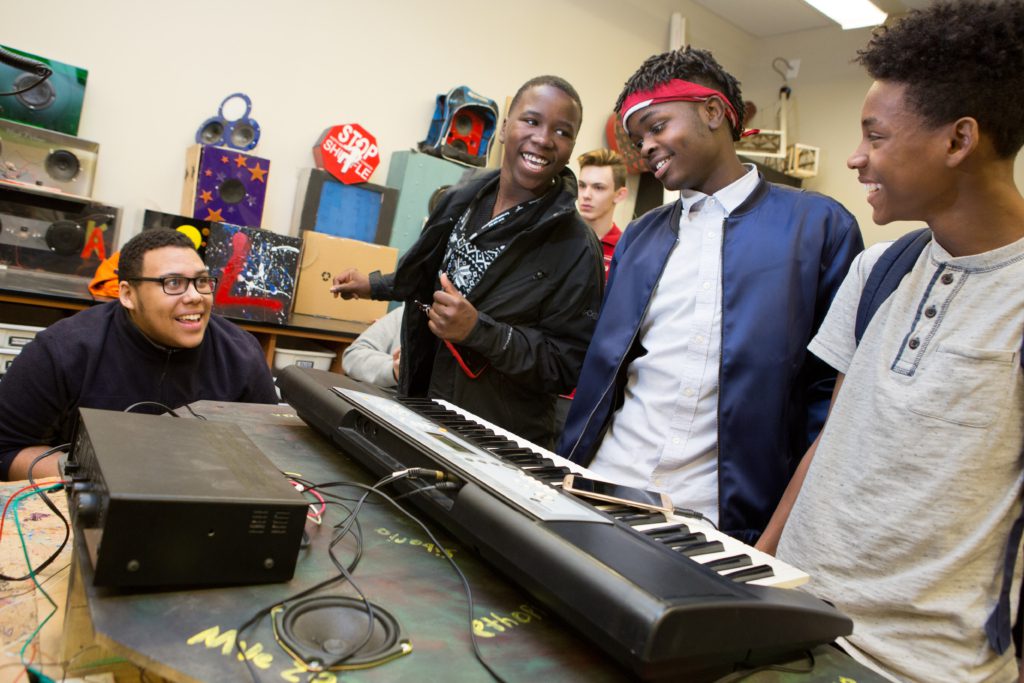 Students standing over a keyboard while talking to a seated teacher