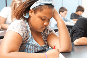 Female black student in a classroom looking down and writing on paper on her desk