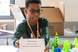A young male black student sitting at a table with his science project