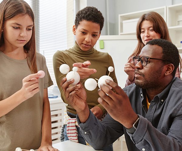 A black male teacher demonstrating a model of planets to three, multiracial female students