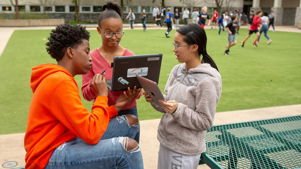 Three middle school students, one male and two female, discussing while holding laptops and tablets outdoors while other students play in the background