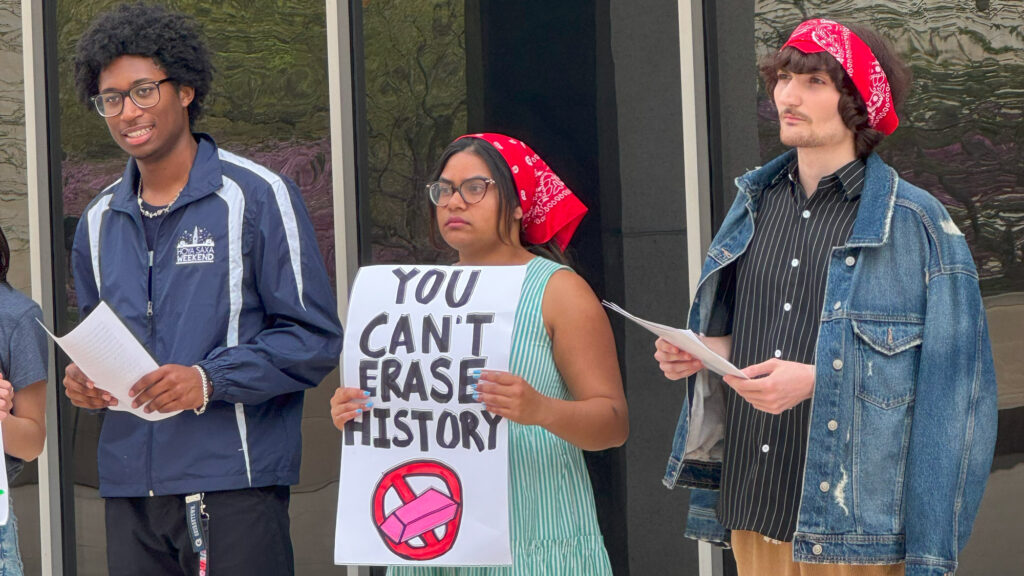 Three students at a protest, two holding paper and one in the middle with a sign that says You Can't Erase History