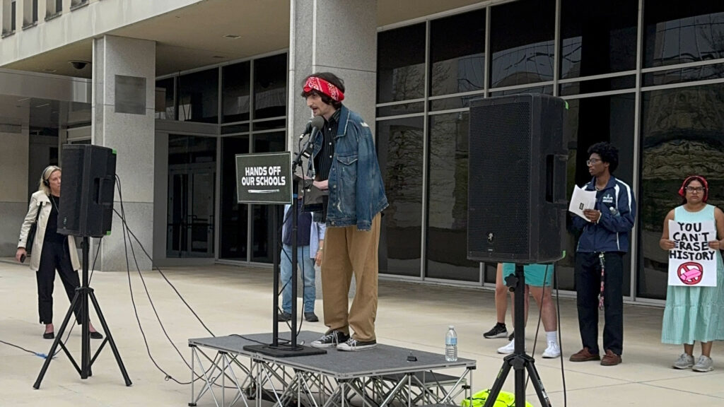 A student wearing a red bandanna speaking on a podium with audio speakers next to a sign that says Hands Off Our Schools