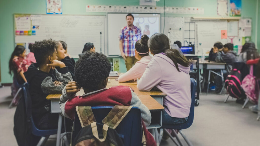 A middle school classroom with young students sitting on a table listening to a male teacher standing in front of a white board