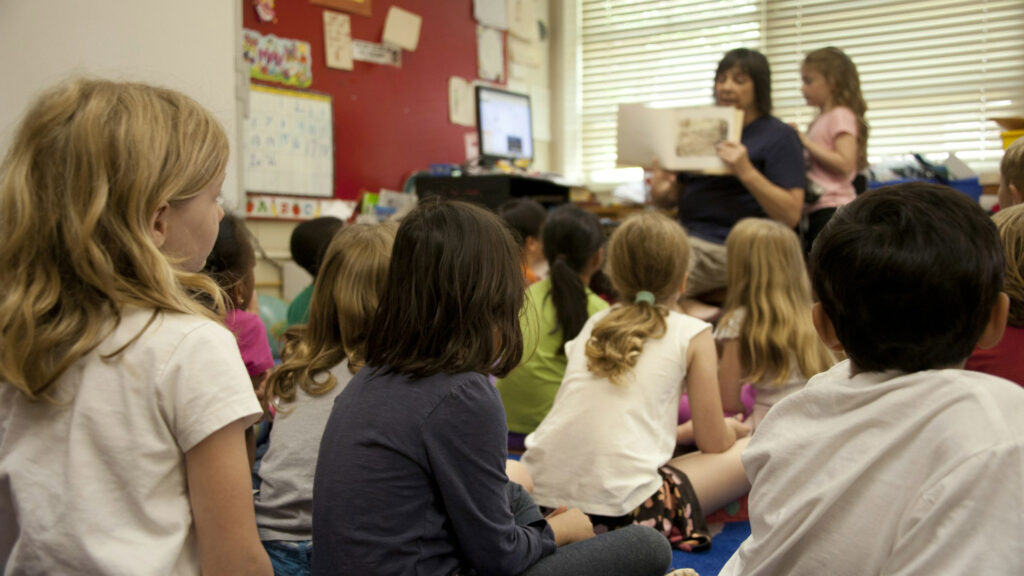 A female teacher reading a book in front of a group of seated young students in a classroom
