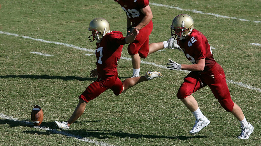 College football player prepares to kick the ball to start the game. His teammates prepare to accompany him downfield.