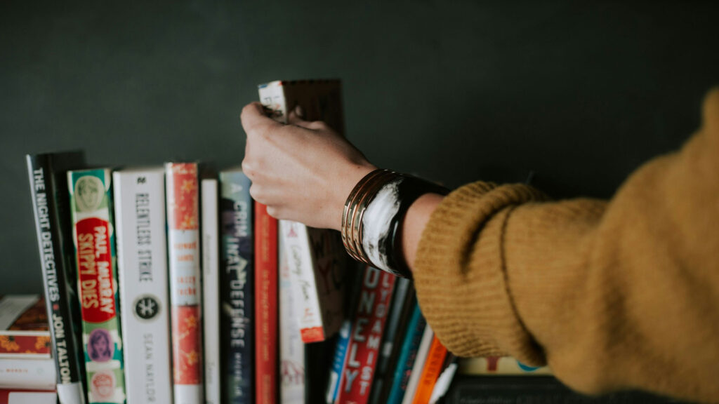 A close up of a hand wearing bracelets reaching out to pick up a paper back book from a row of books