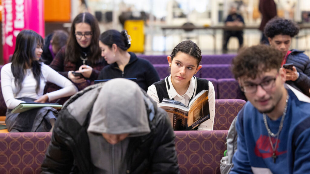 Hispanic female student looks pensive while holding a book in a crowded sitting area on a college campus.