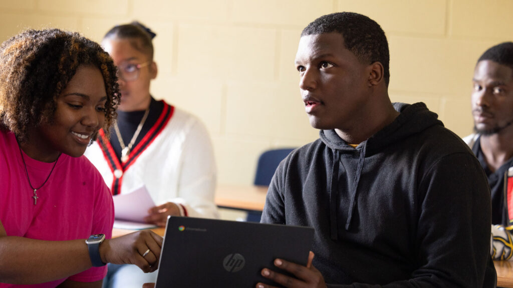 Black female student pointing to a computer held by a Black male student looking ahead during a class on a college campus.