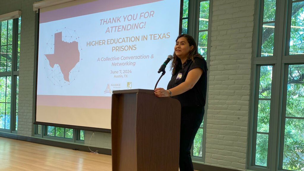 A woman speaking to a microphone on a podium onstage. In the background is a screen that says Thank you for attending! Higher Education in Texas Prisons, A Collective Conversation and Networking
