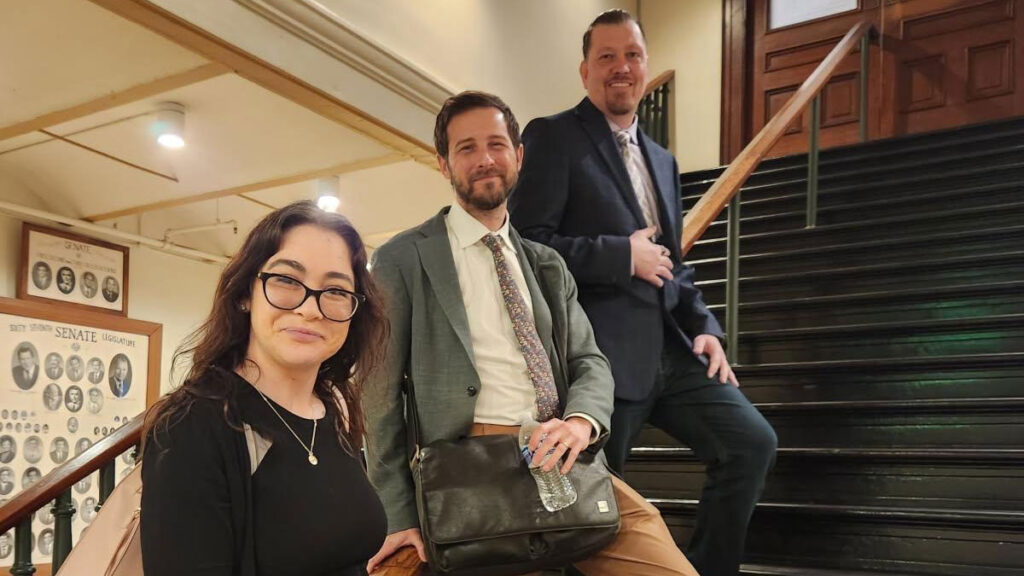 Three people posing on the stairs inside a building. A woman is in front, a man wearing a tie and carrying a shoulder bag is in the middle. On top is a man wearing a suite and tie