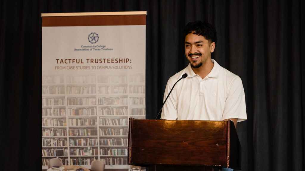 A male student wearing a white shirt speaking onstage at a podium next to a sign with the label Tactful Trusteeship: From Case Studies to Campus Solutions