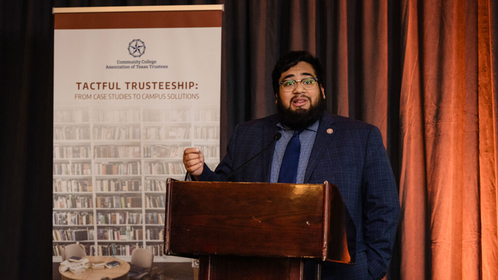 A male student wearing a blue suit and tie speaking onstage at a podium next to a sign with the label Tactful Trusteeship: From Case Studies to Campus Solutions