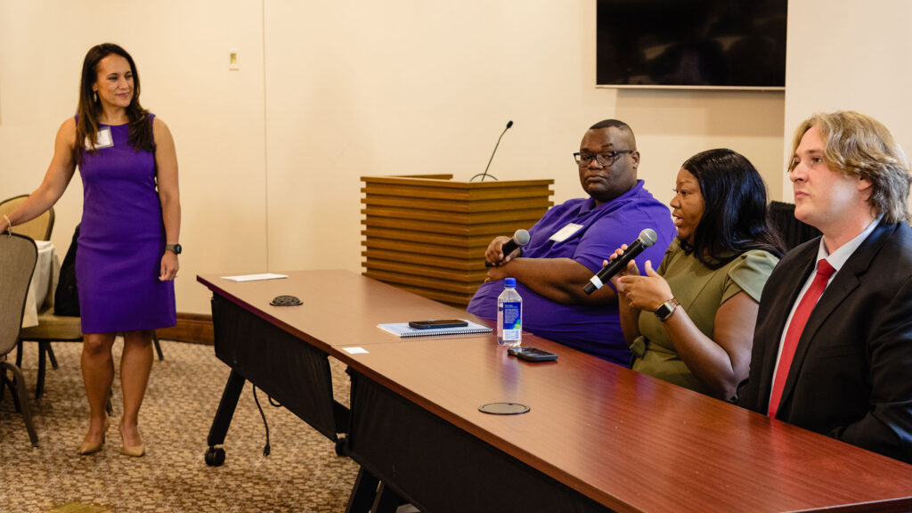 A woman wearing a purple dress stands on the left of a group of three people seated at a table. Two of the seated people hold microphones and the woman at the center is speaking to it