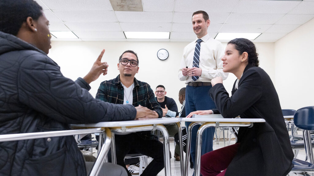 Multiethnic group of students talking with instructor in a classroom on a college campus.