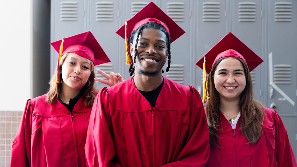Two Hispanic female students and a Black male student in graduation cap and gown posing for a portrait before a graduation ceremony on a college campus.