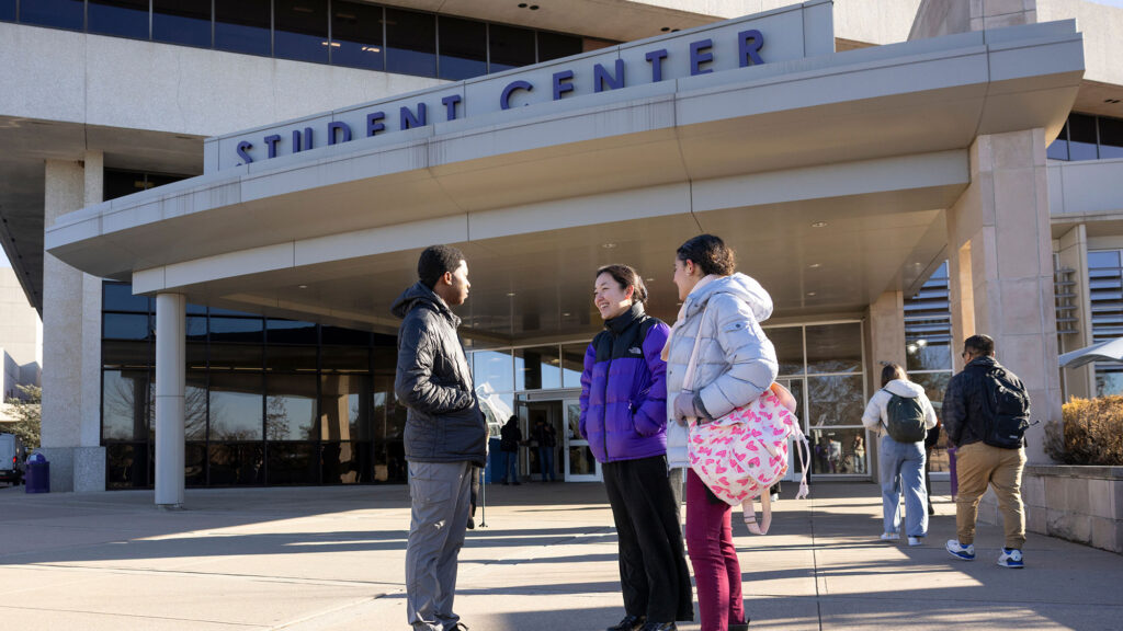 Multiethnic group of students talking together outside of a building labeled "Student Center" on a college campus.