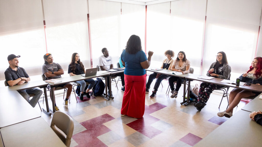 Black female instructor speaking to a multiracial group of students during a class in a sunny classroom on a college campus
