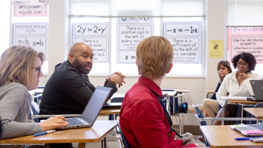 Teachers sitting on desks and and talking in a meeting in a classroom