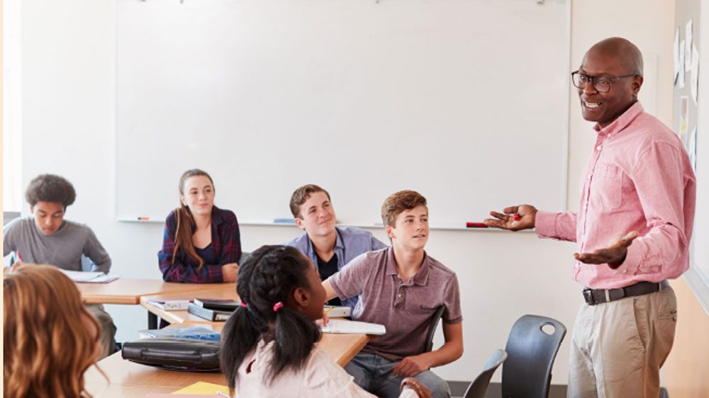 A teacher standing in front or a classroom teaching seated high school students