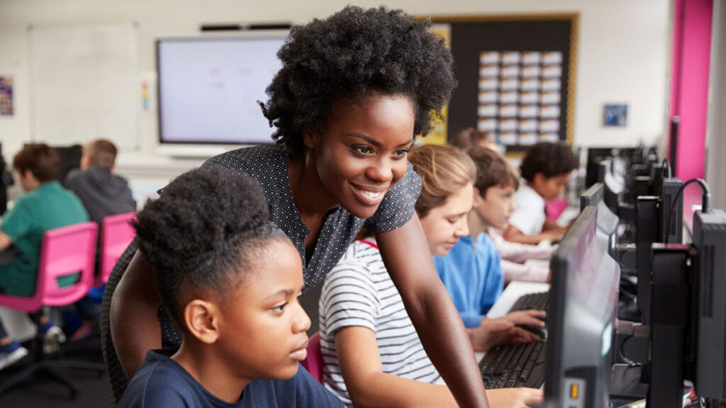 A standing female teacher assisting a seated young female student in front of a computer screen